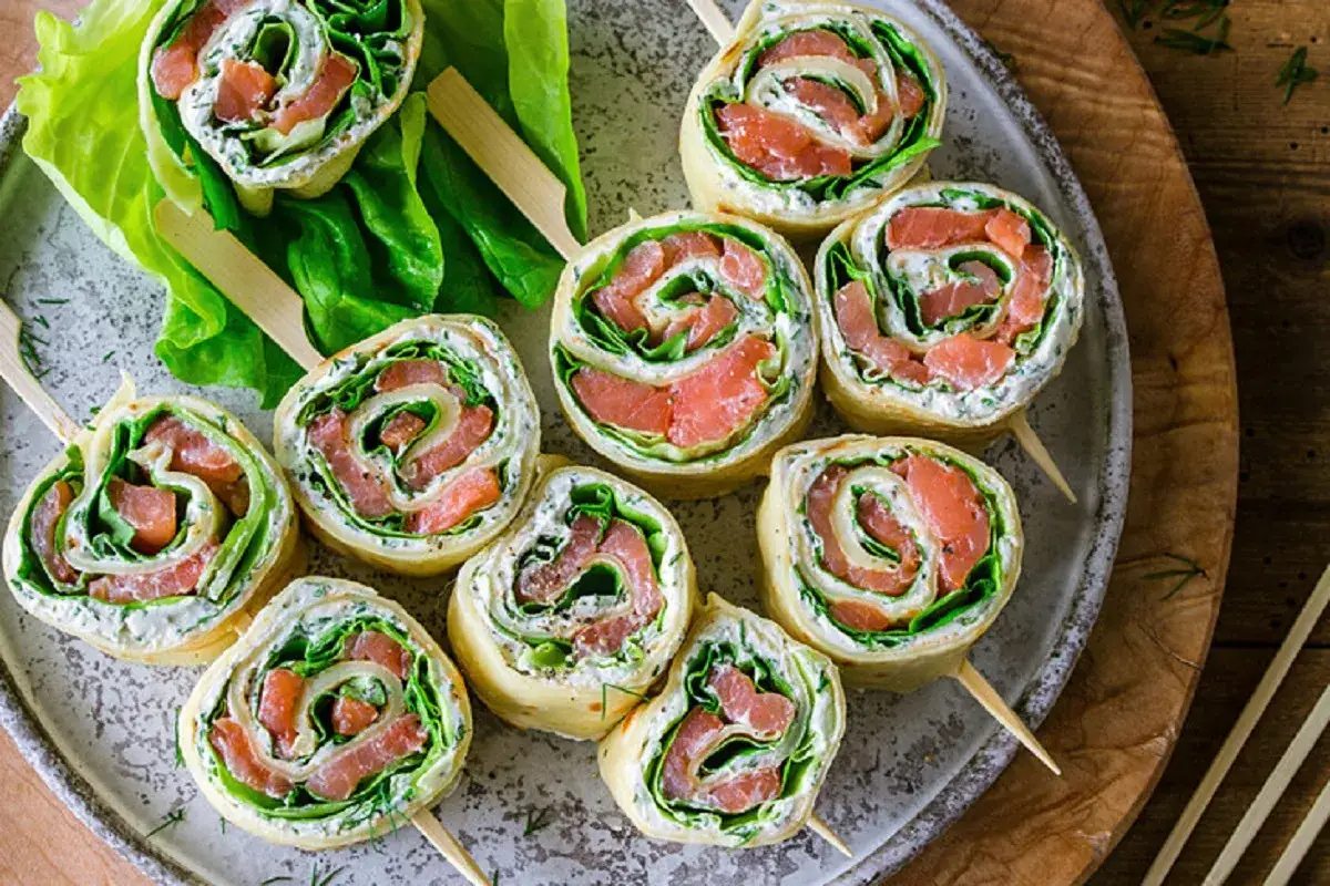 Roladki z tortilli z łososiem, kremowym serkiem i świeżą sałatą, podane na talerzu z liśćmi sałaty.