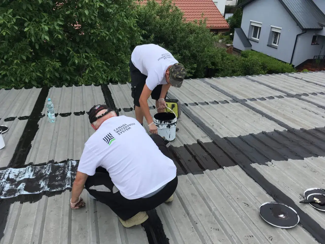 Dw&oacute;ch mężczyzn pracuje na dachu z blachy, uszczelniając go. Pokazują, jak załatać dziurę w dachu z blachy, nakładając czarną masę uszczelniającą.