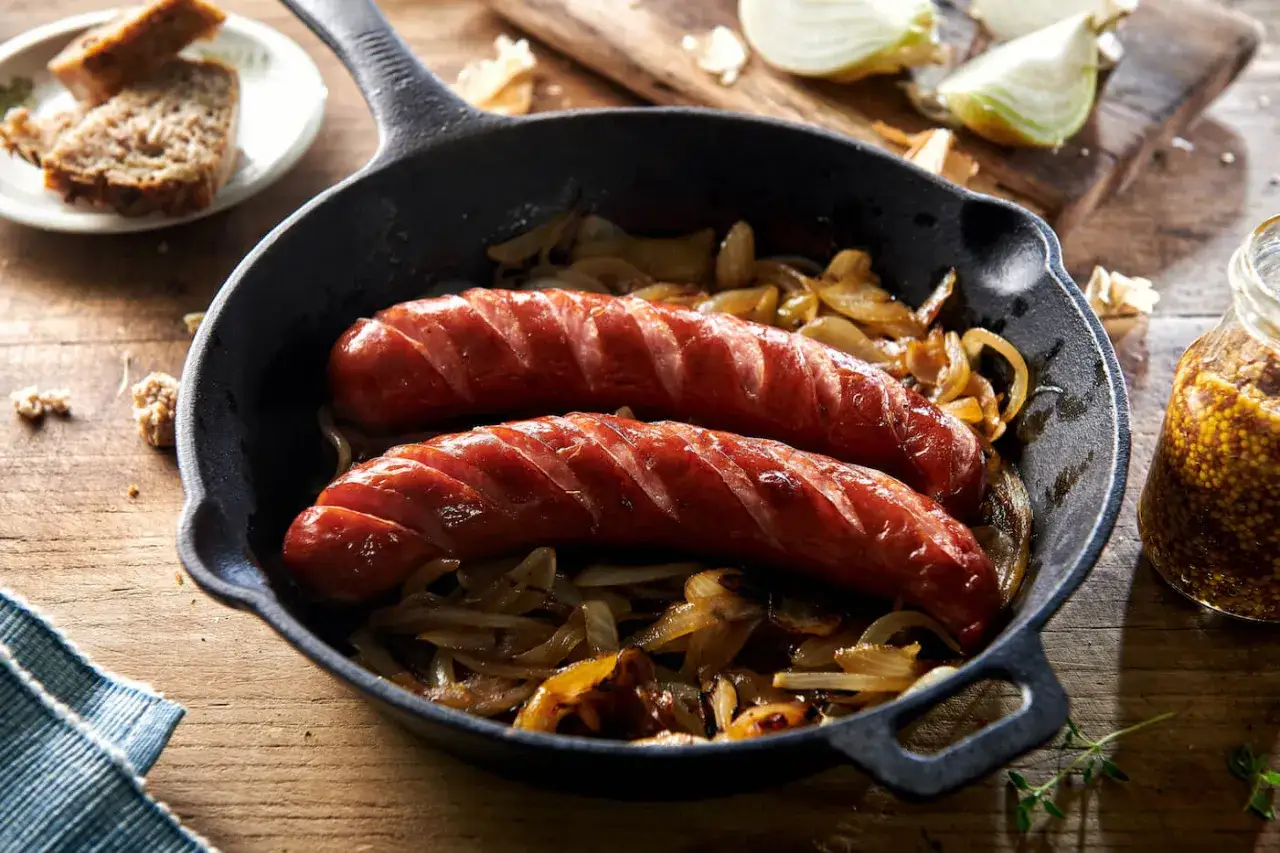 Dwie soczyste kiełbaski z cebulą skwierczące na patelni. Obok kromki chleba i słoiczek musztardy.