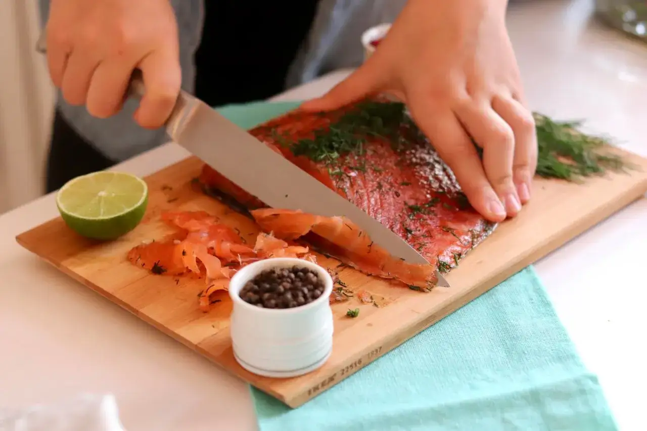 Krojenie wędzonego łososia, który jest częścią marynaty do lososia, na desce z koprem, limonką i kaparami.