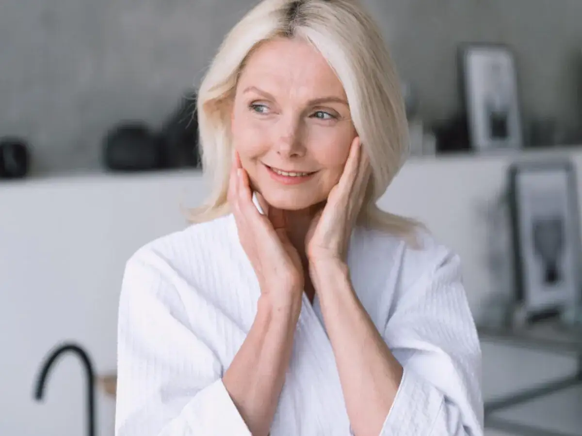 Uśmiechnięta blondynka w szlafroku, dotykająca delikatnie twarzy. Idealny moment na rozważenie, jakie zabiegi na twarz dla 50 latki pomogą zachować młodość.