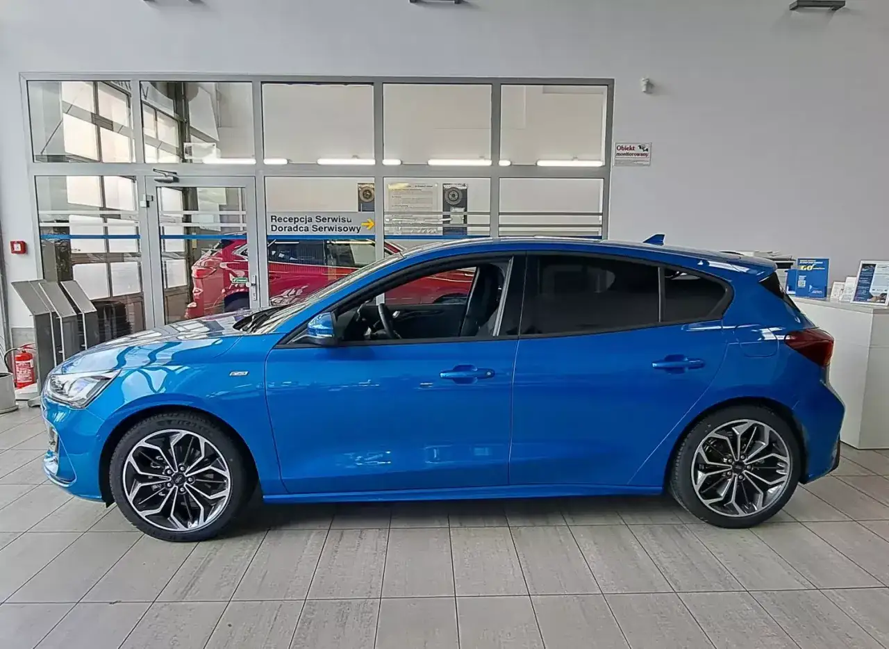 Błękitny Ford Focus 2025 z przyciemnianymi szybami i efektownymi felgami stoi w salonie samochodowym.