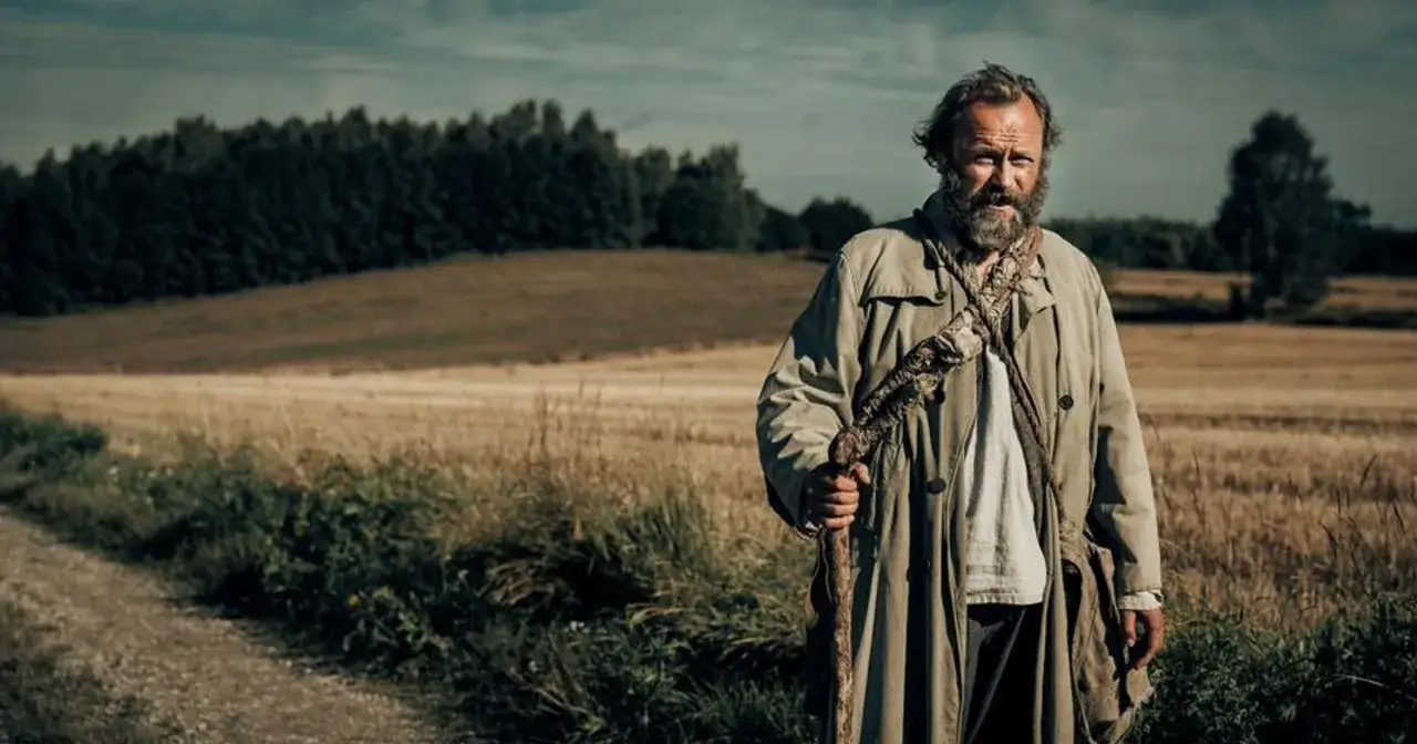 Znachor wędruje przez pola, gdzie rozgrywa się jego historia. W ręku trzyma kostur, a na ramieniu przewieszoną torbę.
