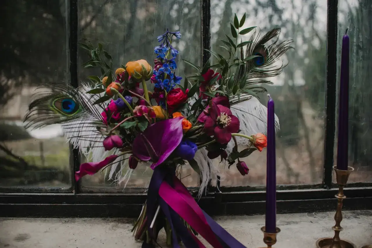 Bogaty bukiet z pawimi pi&oacute;rami i fioletowymi świecami. Kolory kwiat&oacute;w znaczenie: czerwień to namiętność, niebieski to spok&oacute;j, a ż&oacute;łty to radość.