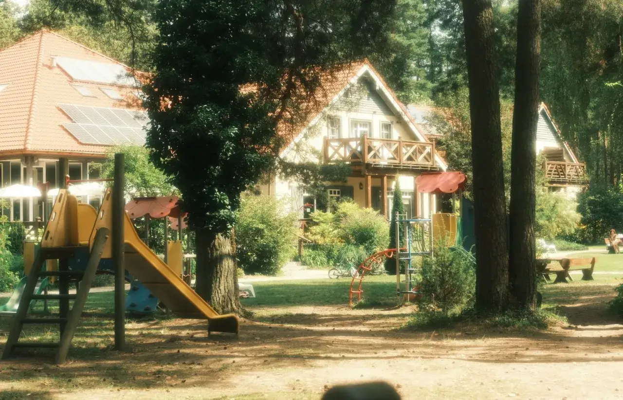 Dzieci bawią się na placu zabaw w otoczeniu zieleni, a w tle widać domki z panelami słonecznymi. Idealne miejsce na rodzinne wakacje mazury all inclusive.