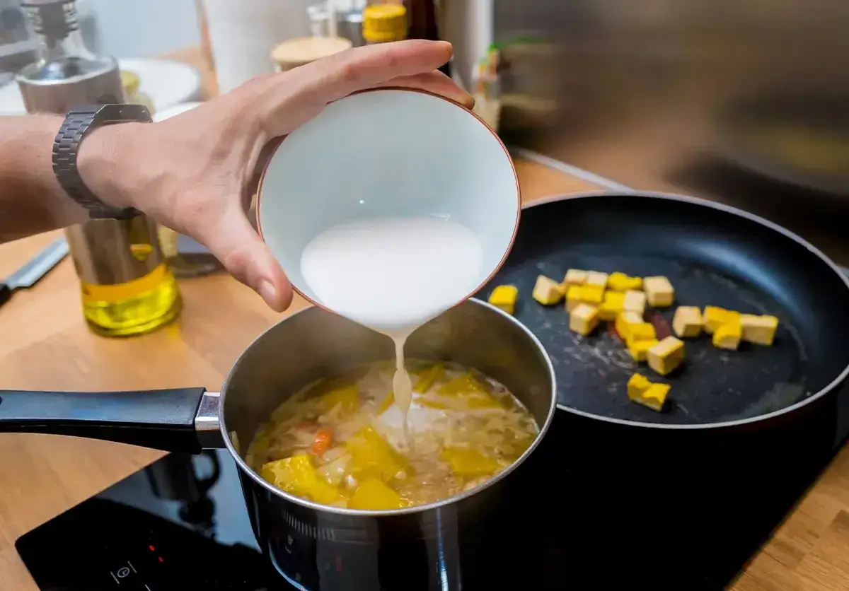 Ręka wylewa białą zawiesinę do garnka z warzywami, by zagęścić zupę gulaszową. Obok smaży się żółte tofu.