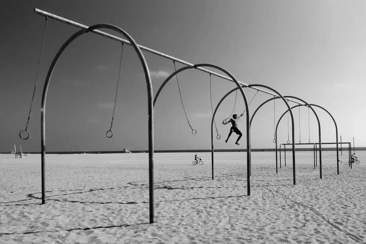 Kompozycja dynamiczna fotografia: mężczyzna ćwiczy na kółkach gimnastycznych na plaży, w tle rowerzysta i ocean.