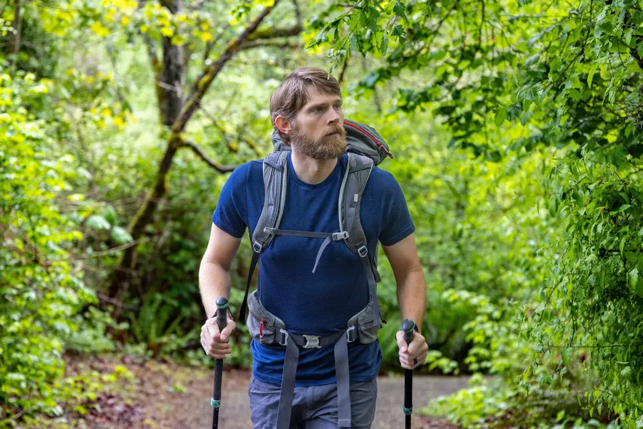 Mężczyzna z brodą wędruje po lesie, ubrany w koszulkę z wełny z merynosa i plecak.
