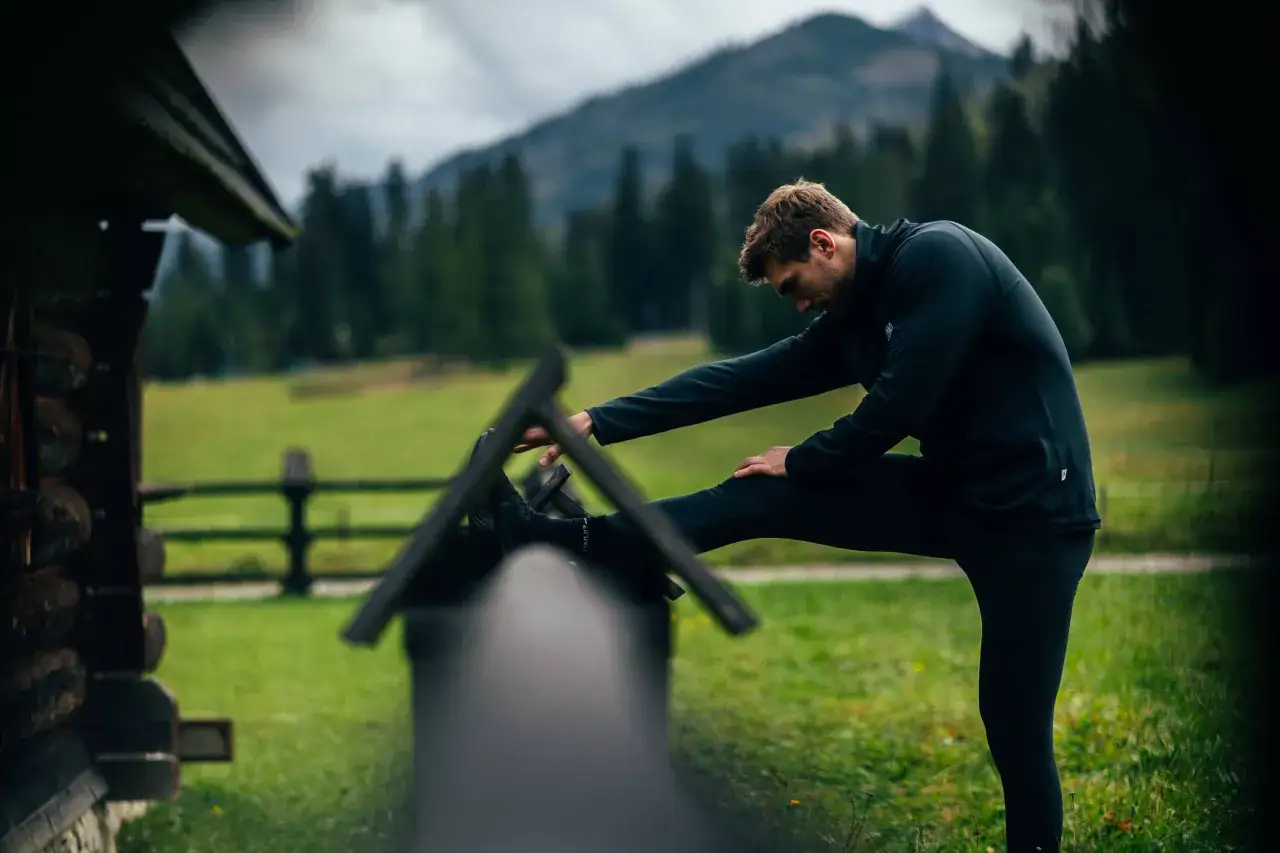 Mężczyzna w czarnym stroju rozciąga nogę przed drewnianą chatką, przygotowując się do biegu anglosaskiego w g&oacute;rskim krajobrazie.