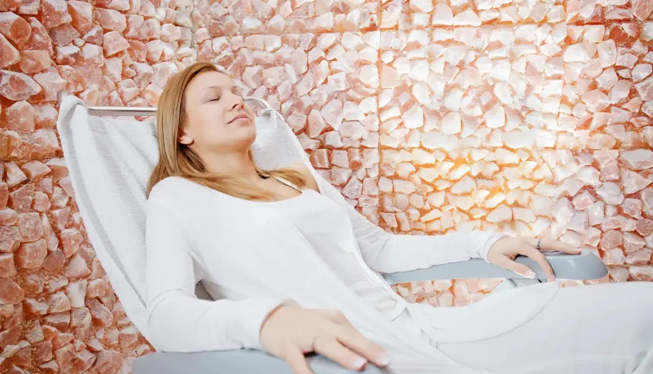 Kobieta w białej bluzce odpoczywa w fotelu, wdychając powietrze z solą do inhalacji. Tło tworzy ściana z r&oacute;żowych kryształ&oacute;w soli.
