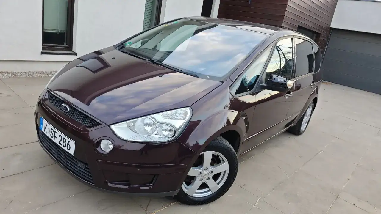 Ciemnobordowy Ford Galaxy lśni w słońcu przed nowoczesnym domem.