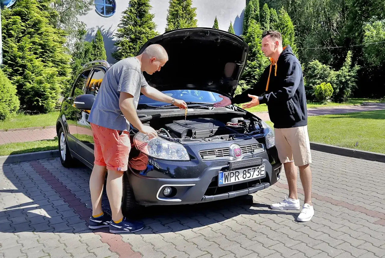 Dwóch mężczyzn przy otwartej masce czarnego auta. Jeden sprawdza poziom oleju, drugi gestykuluje, jakby omawiał stan techniczny. To kluczowe przy negocjowaniu ceny auta.