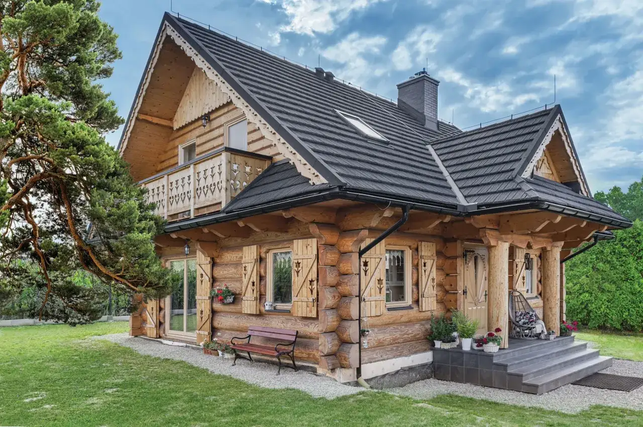 Drewniany dom z okiennicami, otoczony zielenią. Na werandzie huśtawka, przed domem ławka.