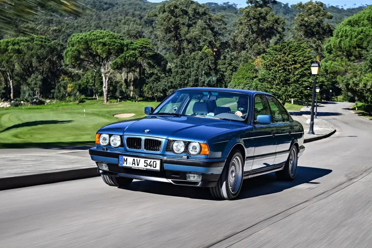 Niebieskie BMW E34 z lat 90. sunie po krętej drodze, mijając malownicze pole golfowe.