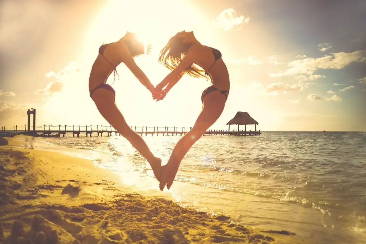 Dwie kobiety w bikini tworzą serce na plaży podczas babskiego wypadu. Słońce zachodzi nad oceanem, a w tle widać pomost.