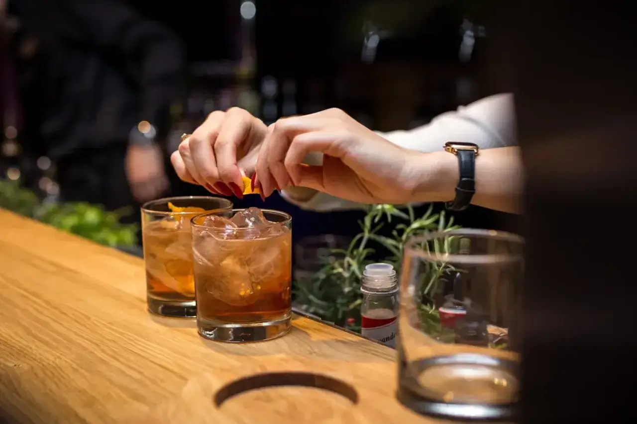 Dłonie z czerwonymi paznokciami dodają skórkę pomarańczy do drinków z lodem. Idealny sposób, jak pić bourbon.