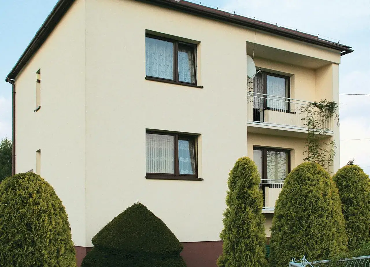 Jasny dom kostka z balkonem i brązowymi oknami. Elewacja czysta, otoczona zielonymi krzewami.