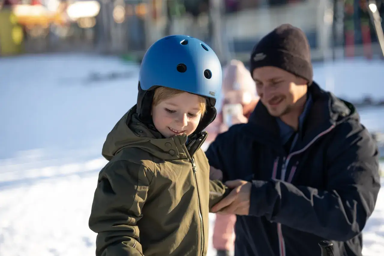 Chłopiec w niebieskim kasku i zielonej kurtce przygotowuje się do jazdy na nartach. Tata pomaga mu, a w tle widać inne osoby i śnieg. Idealne narty dla 3 latka czekają na pierwsze zjazdy.