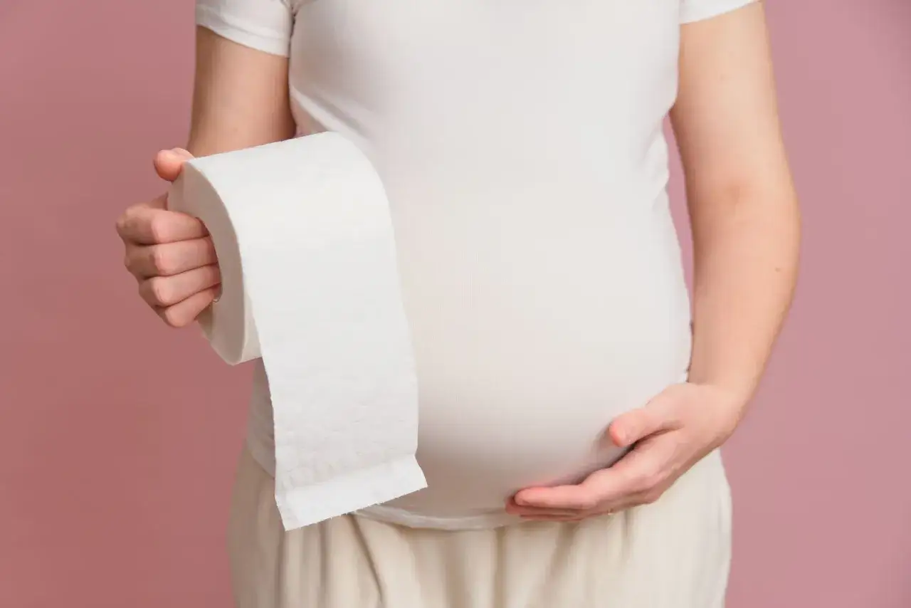 Brzuch kobiety w ciąży, trzymającej rolkę papieru toaletowego. Jelitówka w ciąży to częsty problem.
