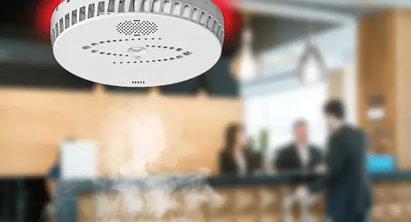 Osoba paląca papierosa w pokoju hotelowym z widocznym czujnikiem dymu