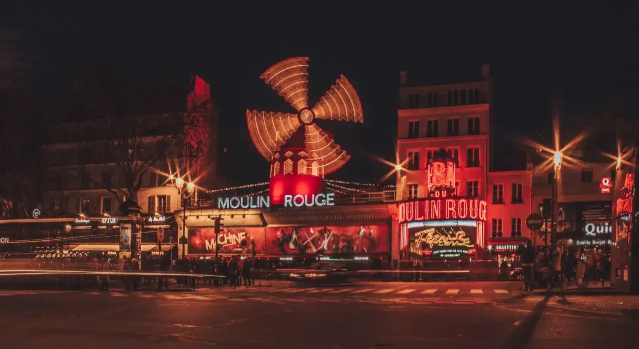 Nocny Paryż rozświetlony neonami kabaretu Moulin Rouge. Charakterystyczny wiatrak wiruje, zapraszając do świata rozrywki.