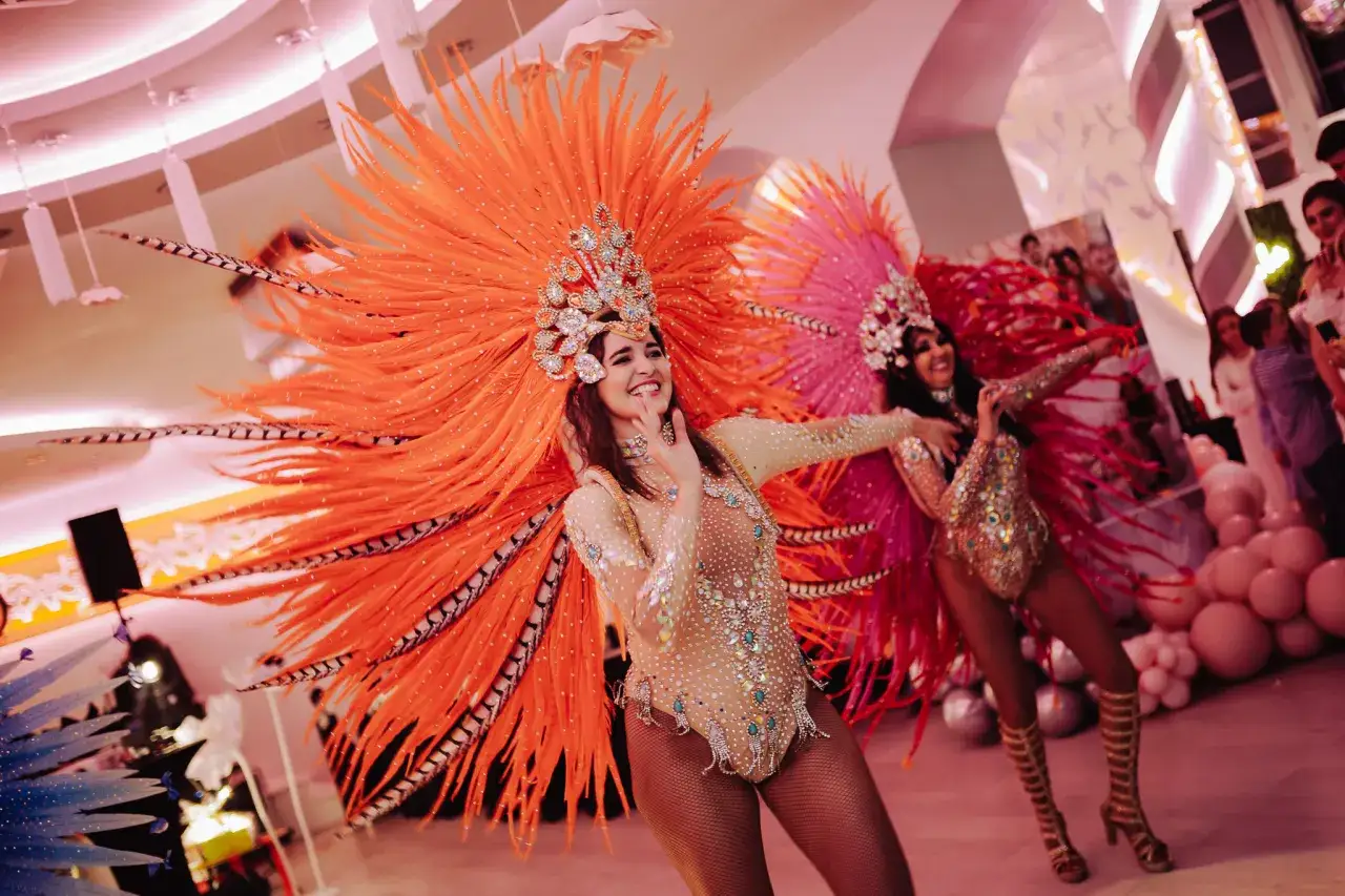 Tancerki w barwnych strojach z pi&oacute;r wykonują piosenki z układem tanecznym. Ich kostiumy zdobią błyszczące kamienie i wysokie buty.