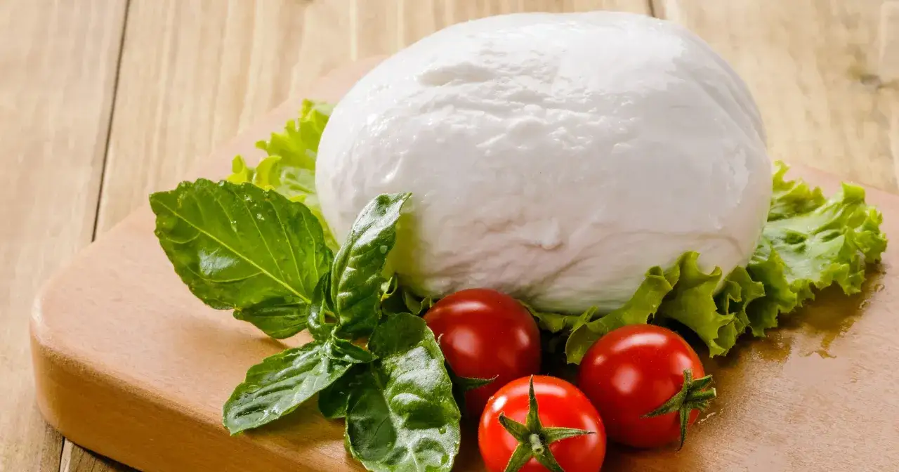 Duża kula białej mozzarelli, pomidorki koktajlowe i liście bazylii na desce. Czy mozzarella jest zdrowa? Tak, w umiarkowanych ilościach.