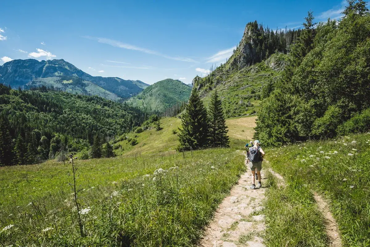 Turyści idą kamienistą ścieżką nad reglami, otoczeni zielonymi wzgórzami i skalistymi szczytami.