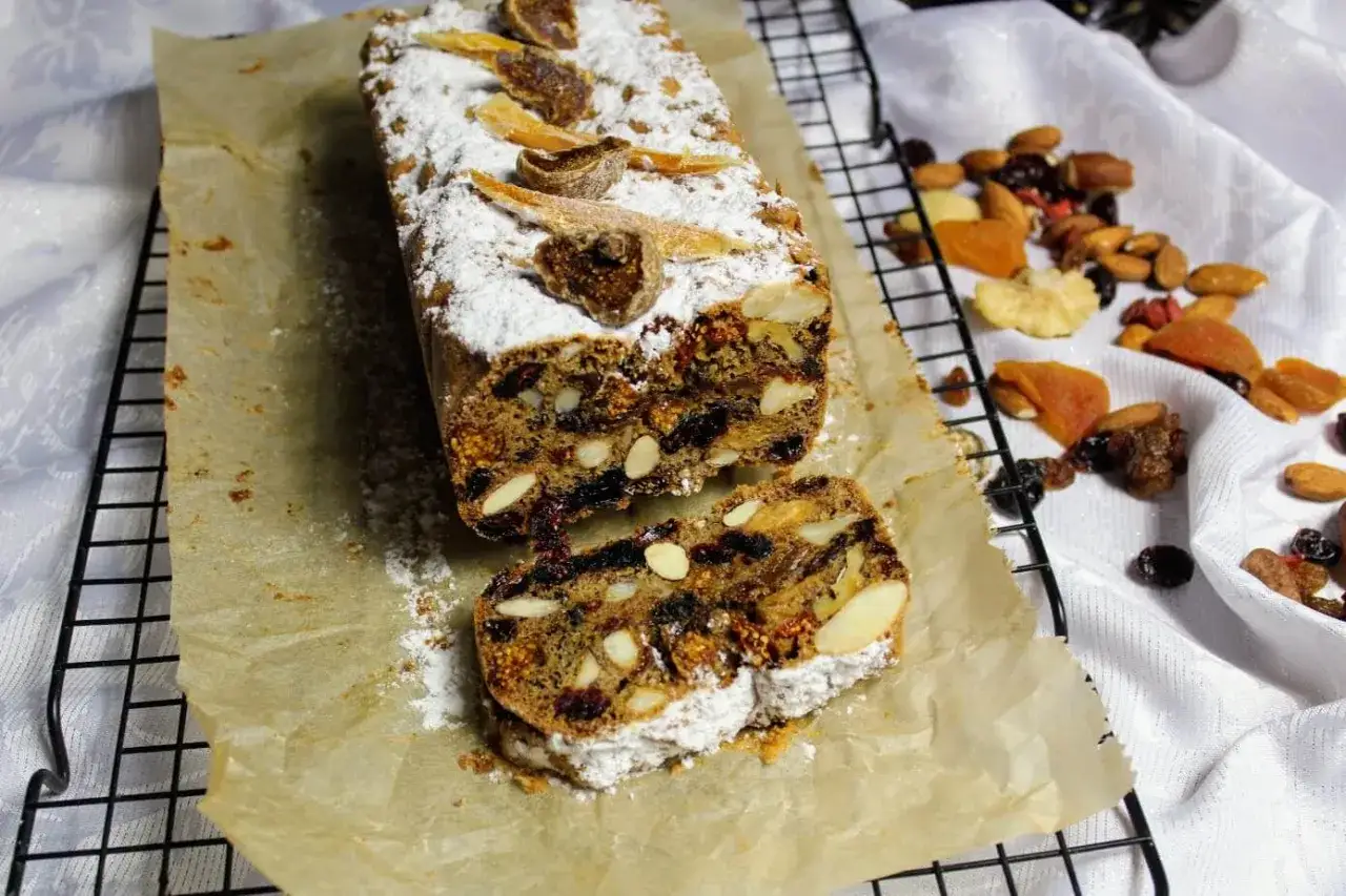 Pyszny keks Ania gotuje, pełen bakalii i migdał&oacute;w, posypany cukrem pudrem. Obok ułożone suszone owoce i orzechy.