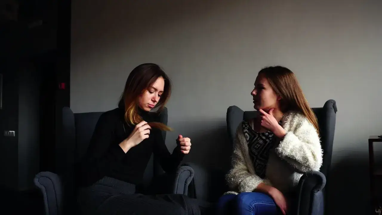 Dwie młode kobiety siedzą w fotelach. Jedna z nich, zamyślona, bawi się kosmykiem włos&oacute;w. Druga, z ręką na szyi, słucha jej uważnie. Może rozmawiają o tym, że są **chętne na dziecko**.