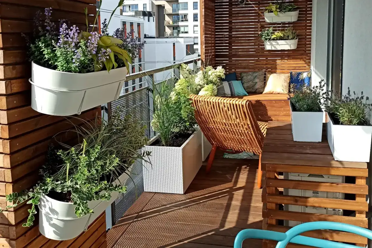 Zielony, kwitnący mały balkon balkon w bloku. Wygodne meble, rattanowy fotel i mnóstwo roślin tworzą oazę spokoju.