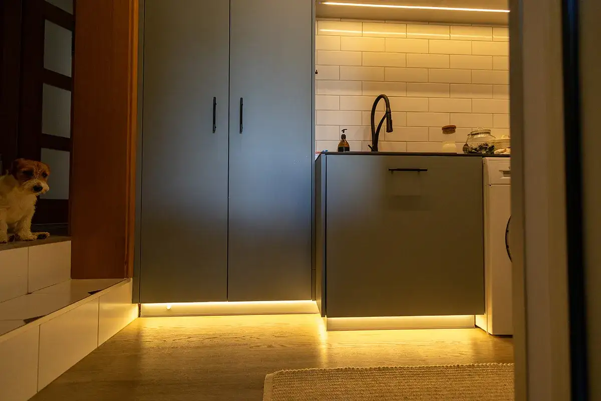 Nowoczesna kuchnia z szarymi frontami i ciepłym oświetleniem cokołu. Obok stoi piesek.