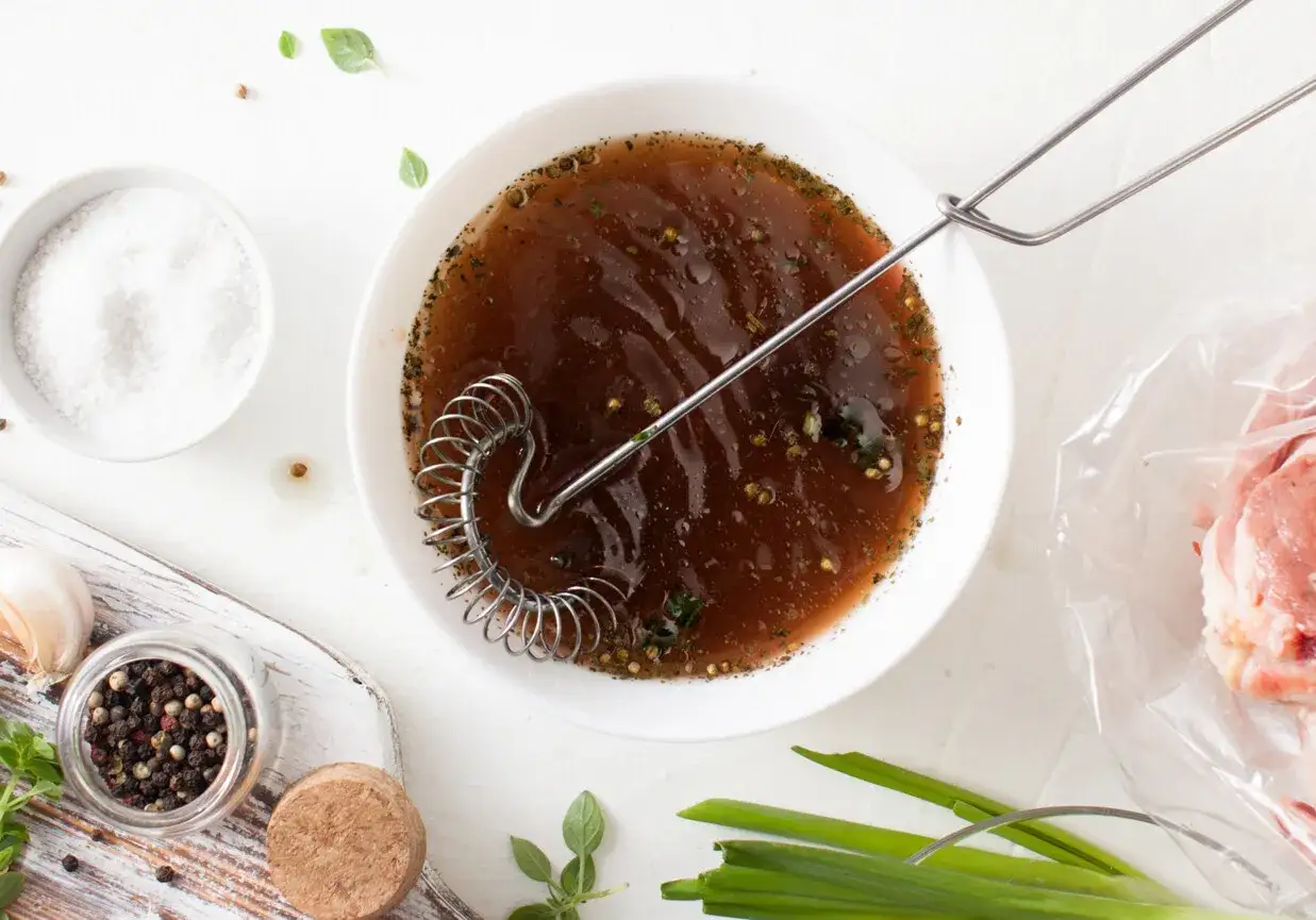 Przygotowanie zalewy do peklowania mięsa. W misce brązowy płyn z przyprawami, obok s&oacute;l, pieprz, czosnek i surowe mięso w folii.