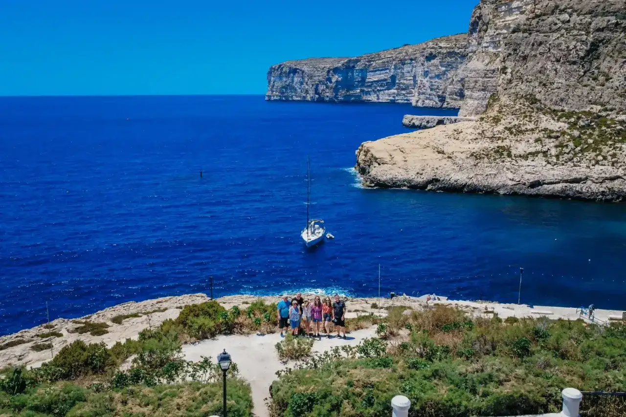 Grupa ludzi na klifie z widokiem na błękitne morze i żagl&oacute;wkę. Malta czy warto? Tak, warto!