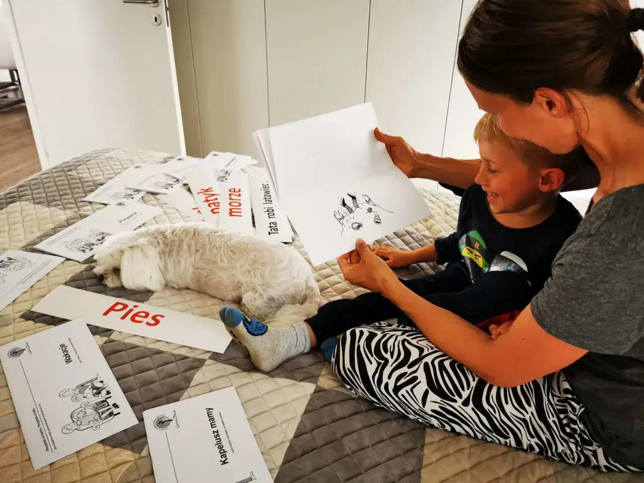 Mama uczy synka czytać, pokazując mu karty ze słowami i obrazkami. Piesek leży obok, a na łóżku leżą materiały do nauki.