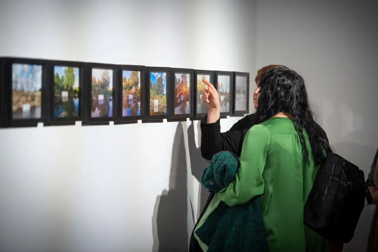 Wystawa Piotra K. Kowalskiego. Dwie kobiety oglądają rzędy małych, czarnych ram z odbiciami drzew i nieba w wodzie.