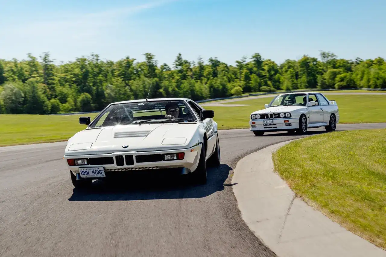 Dwa białe BMW, klasyczne BMW M1 i BMW E30, pędzą po torze wyścigowym w słoneczny dzień.