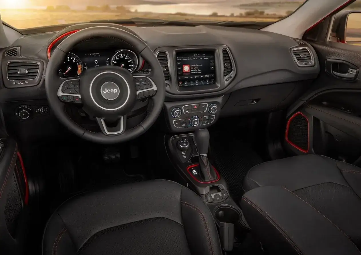 Wnętrze Jeep Compass z czarną tapicerką i czerwonymi akcentami. Widoczna kierownica, deska rozdzielcza z ekranem multimedialnym i dźwignia zmiany biegów.