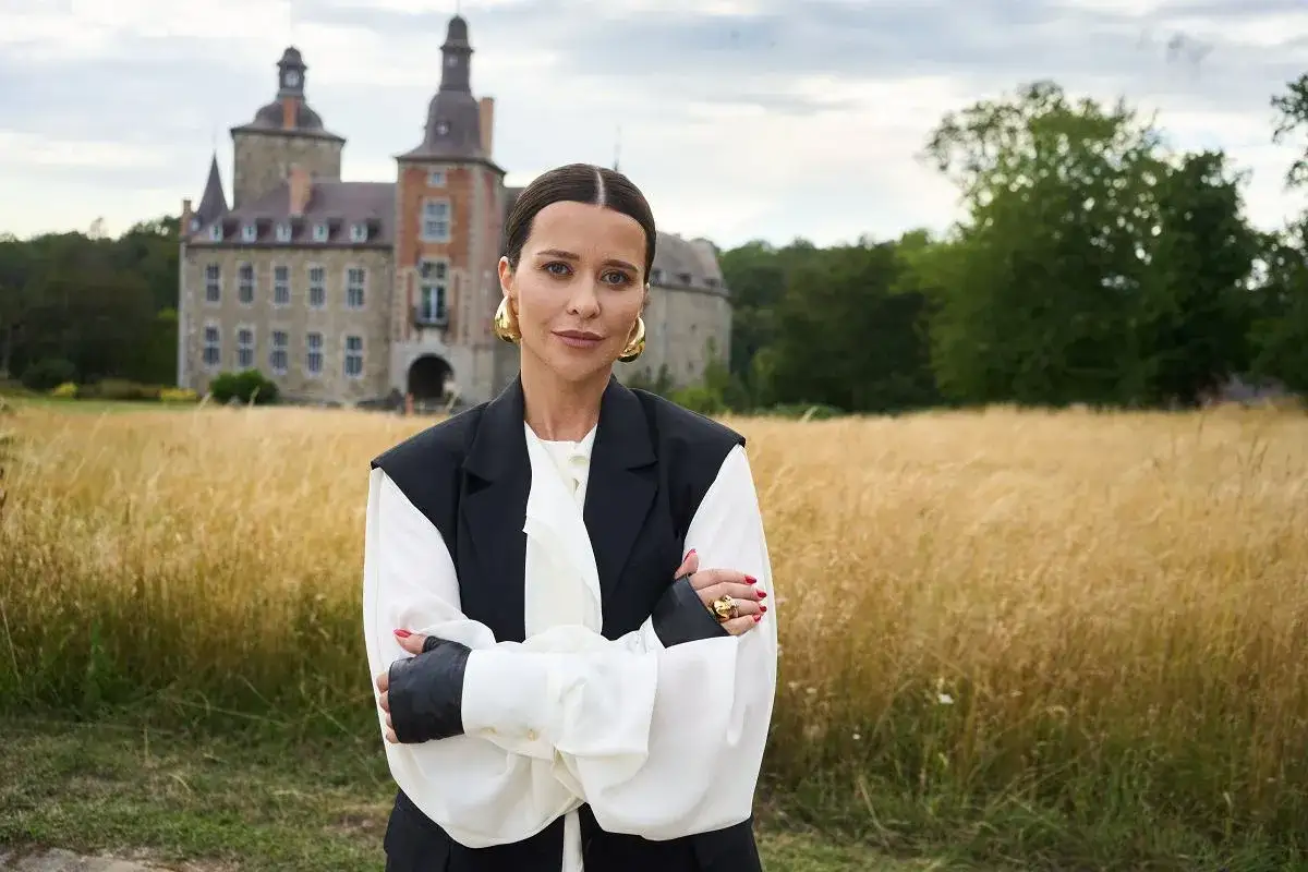 Kobieta w eleganckim stroju, z dużymi złotymi kolczykami, stoi na tle zamku i pola. Czyżby to ona była tą 