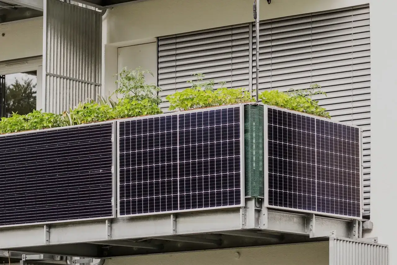 Panele fotowoltaiczne na balkonie, z zielonymi roślinami wyrastającymi ponad nimi. Ekologiczne rozwiązanie dla miejskiego ogrodu.
