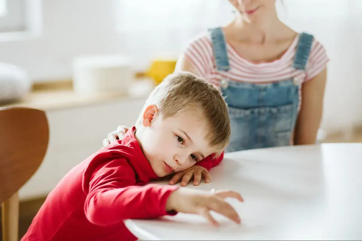 Chłopiec w czerwonej bluzce opiera głowę na stole, jego wzrok błądzi. Mama kładzie mu rękę na ramieniu, wspierając go w walce z problemem z koncentracją u dziecka.