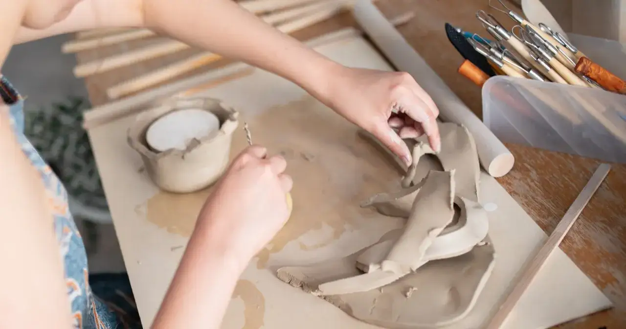 Dłonie formują glinę, pokazując, co można zrobić z gliny: miseczki, figurki i inne dzieła sztuki. Narzędzia ceramiczne czekają na użycie.