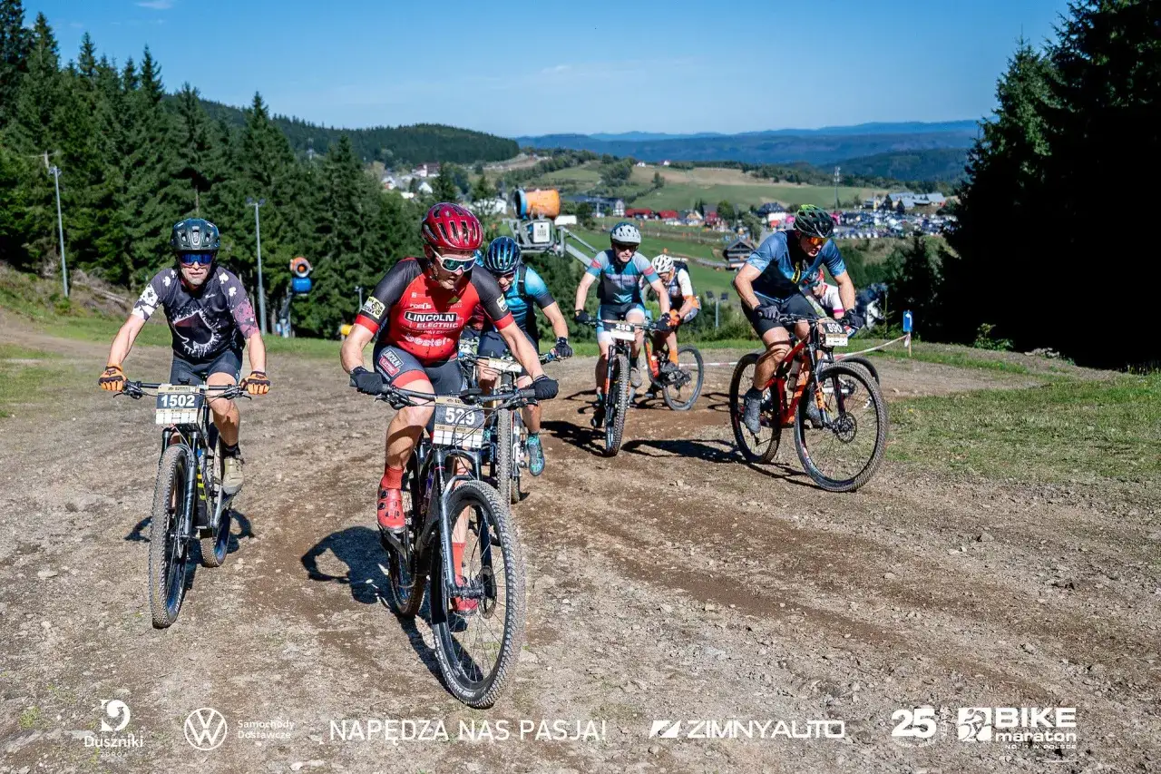 Kolarze ścigają się na górskiej trasie podczas zawodów MTB. Słońce, las i malownicze krajobrazy towarzyszą uczestnikom.