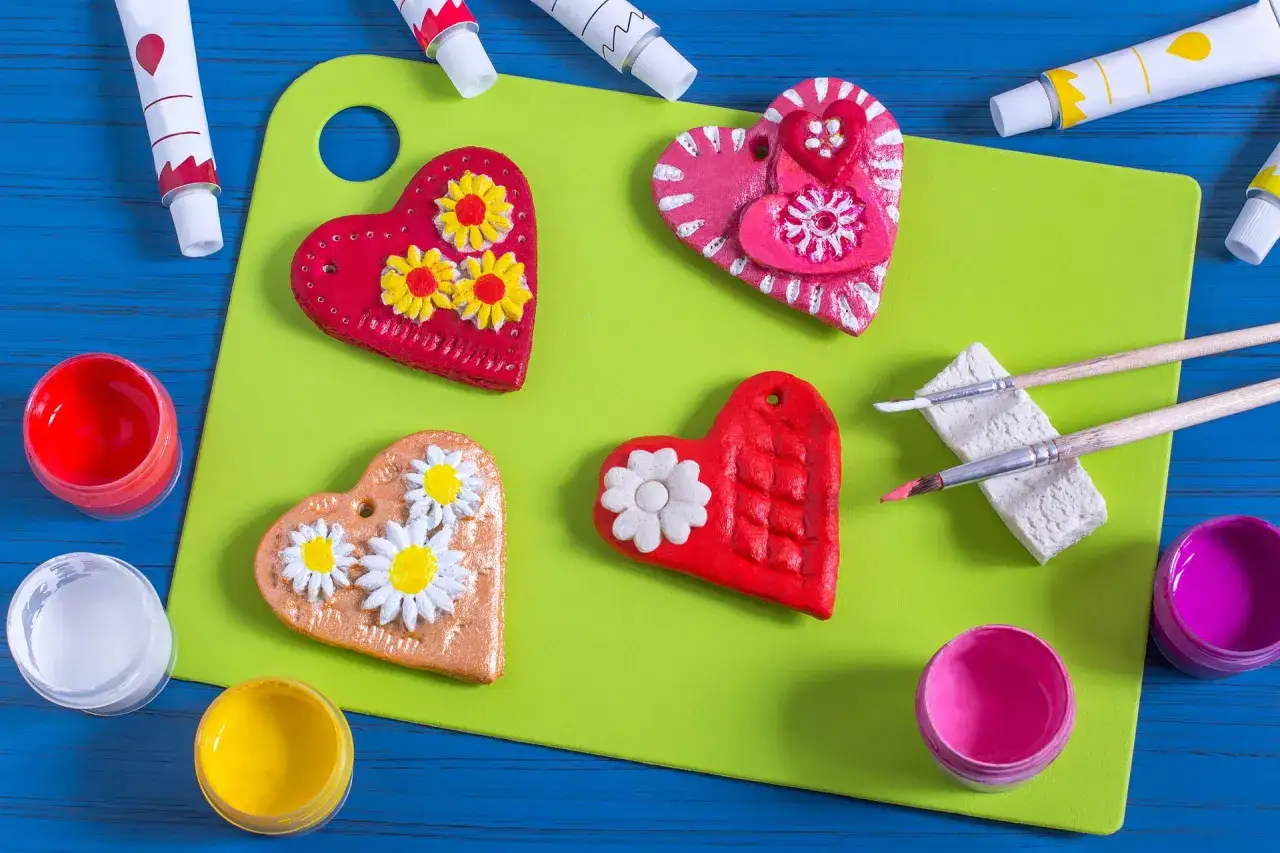 Kolorowe serca z masy solnej, ozdobione kwiatami, czekają na malowanie. Obok farby i pędzle.
