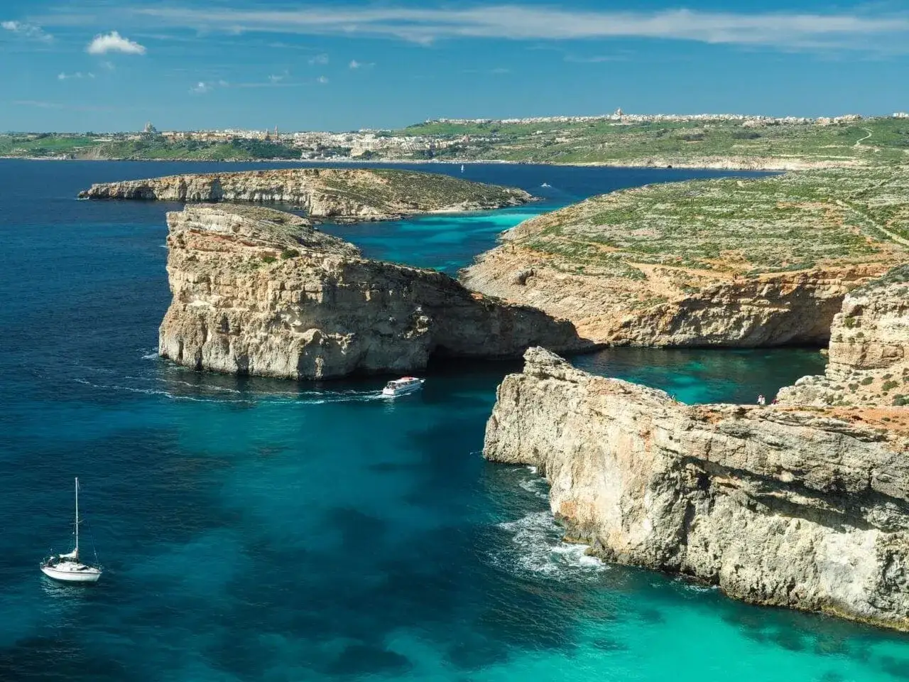 Malta: skaliste wybrzeże z turkusową wodą, łódź i żaglówka. Idealne miejsce, kiedy lecieć na Maltę, by cieszyć się słońcem.