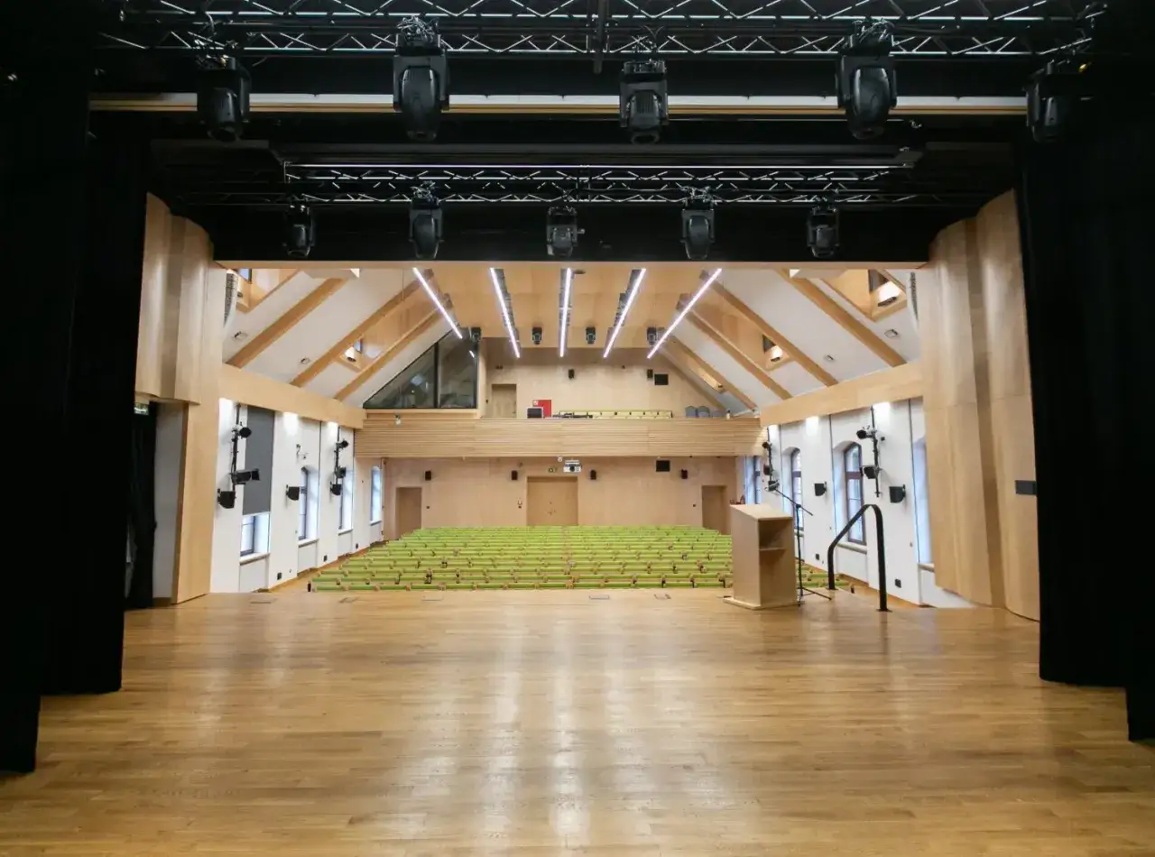 Widok na pustą aulę z drewnianą sceną, rzędami zielonych krzeseł i podestem dla mówcy. To idealne miejsce na wykłady i wydarzenia.