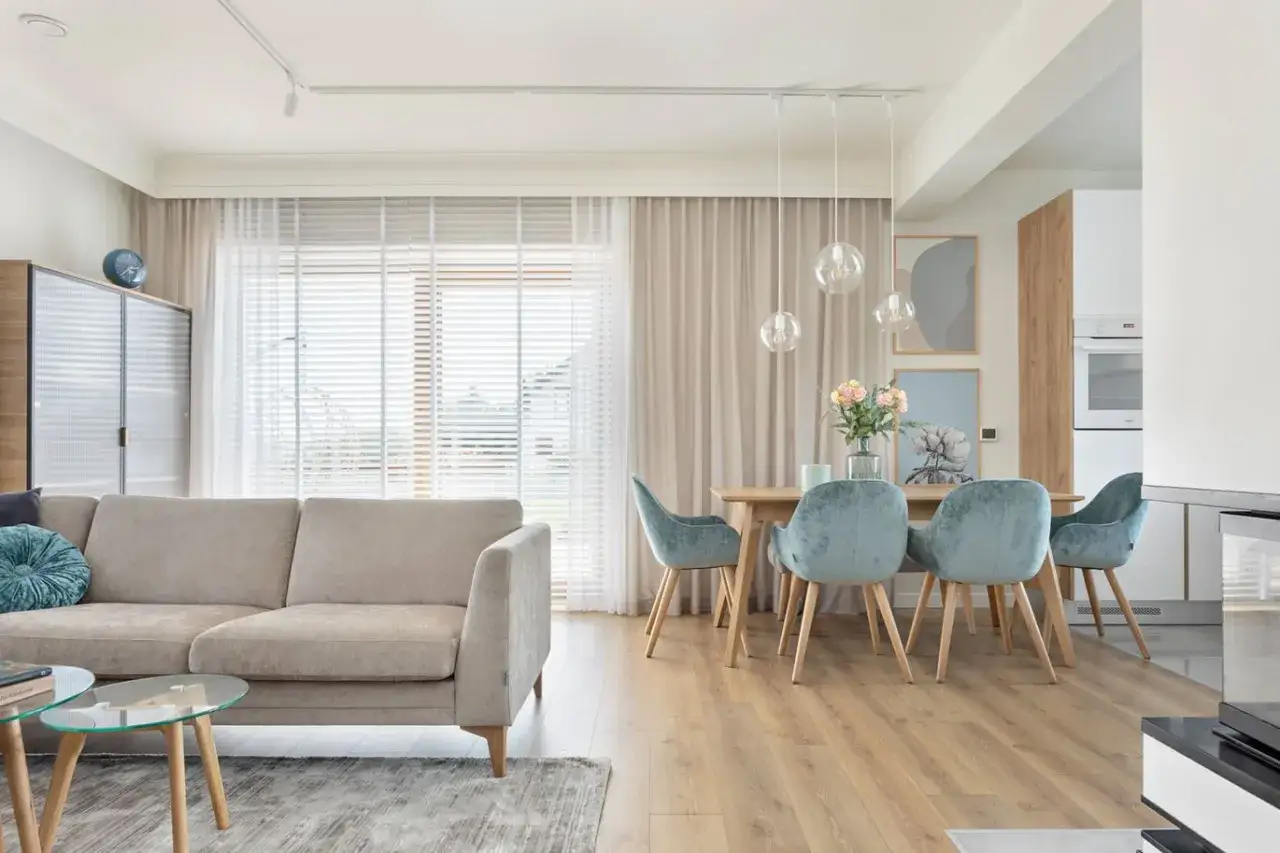 Jasny salon z beżową kanapą i stołem jadalnym. Nowoczesne zasłanianie okien tworzy przytulną atmosferę.