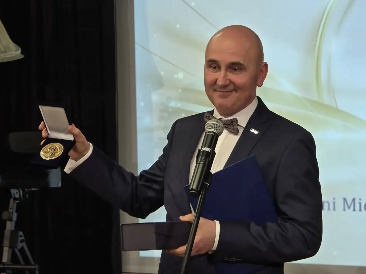 Prezes spółdzielni mieszkaniowej, osoba publiczna, odbiera medal. Uśmiecha się, trzymając w dłoniach medal i dyplom.