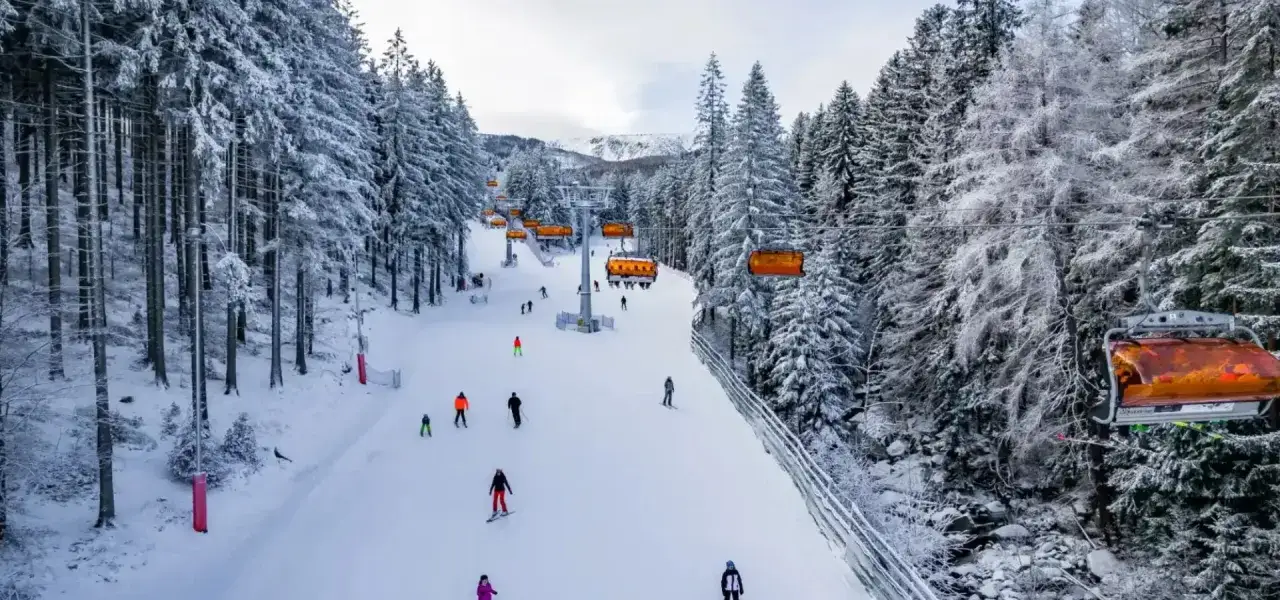 Zimowy dzień na stoku. Wyciąg narciarski Karpacz z pomarańczowymi kanapami w otoczeniu ośnieżonych drzew. Narciarze zjeżdżają po przygotowanej trasie.