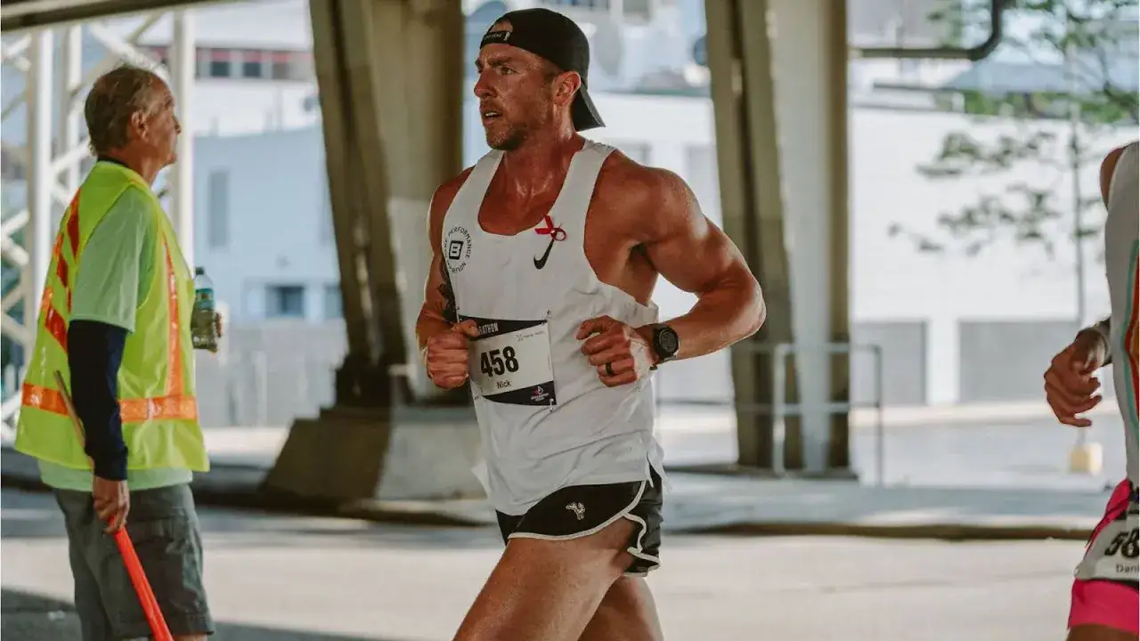 Biegacz Nick z numerem 458, w białej koszulce, biegnie podczas maratonu. Widoczny jest też wolontariusz w kamizelce odblaskowej. To inspiracja, jak przygotować się do maratonu.